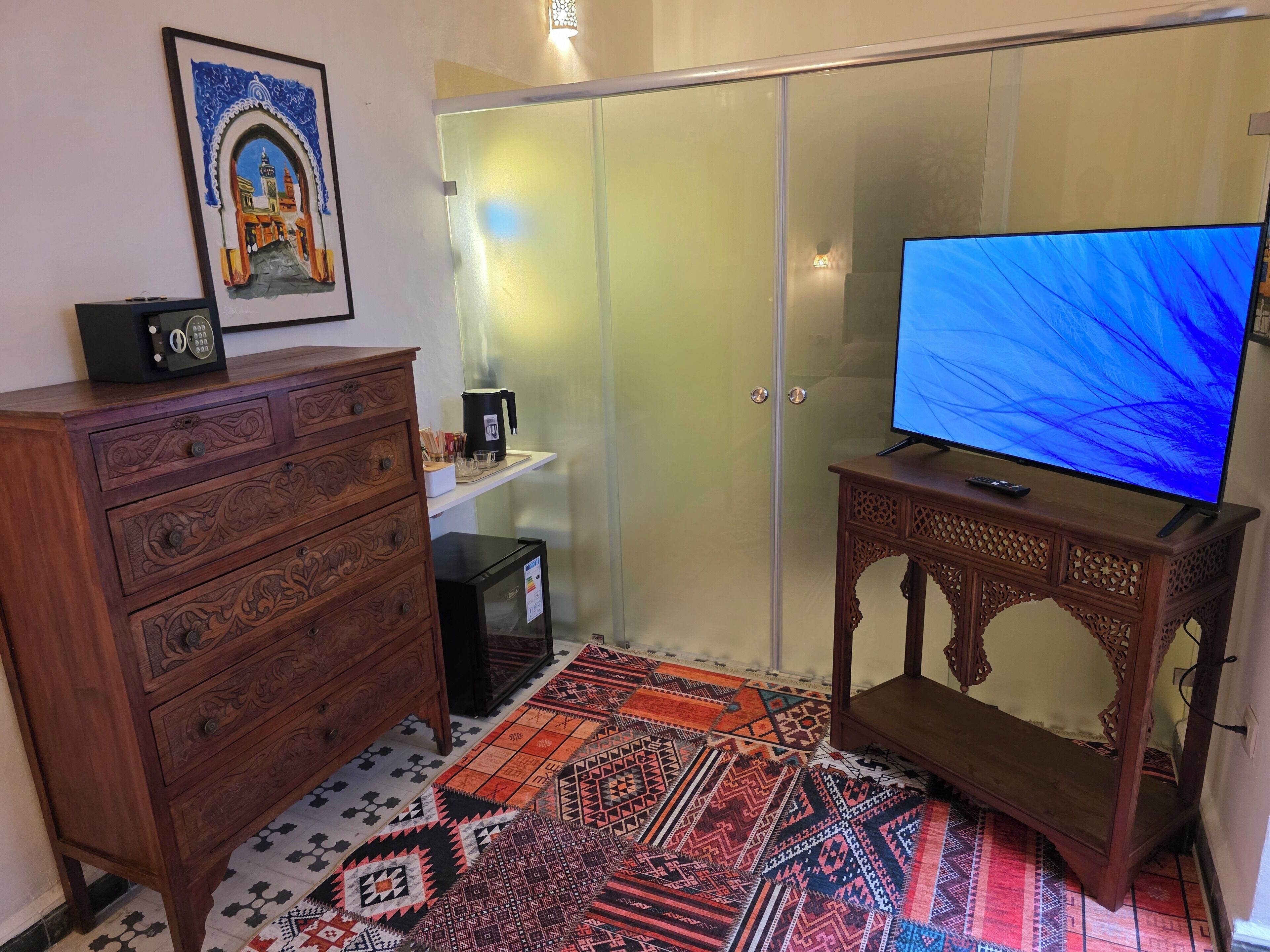 A room with a television on a wooden stand, a small refrigerator, a cabinet with drawers, a patterned rug, and a door with frosted glass panels.