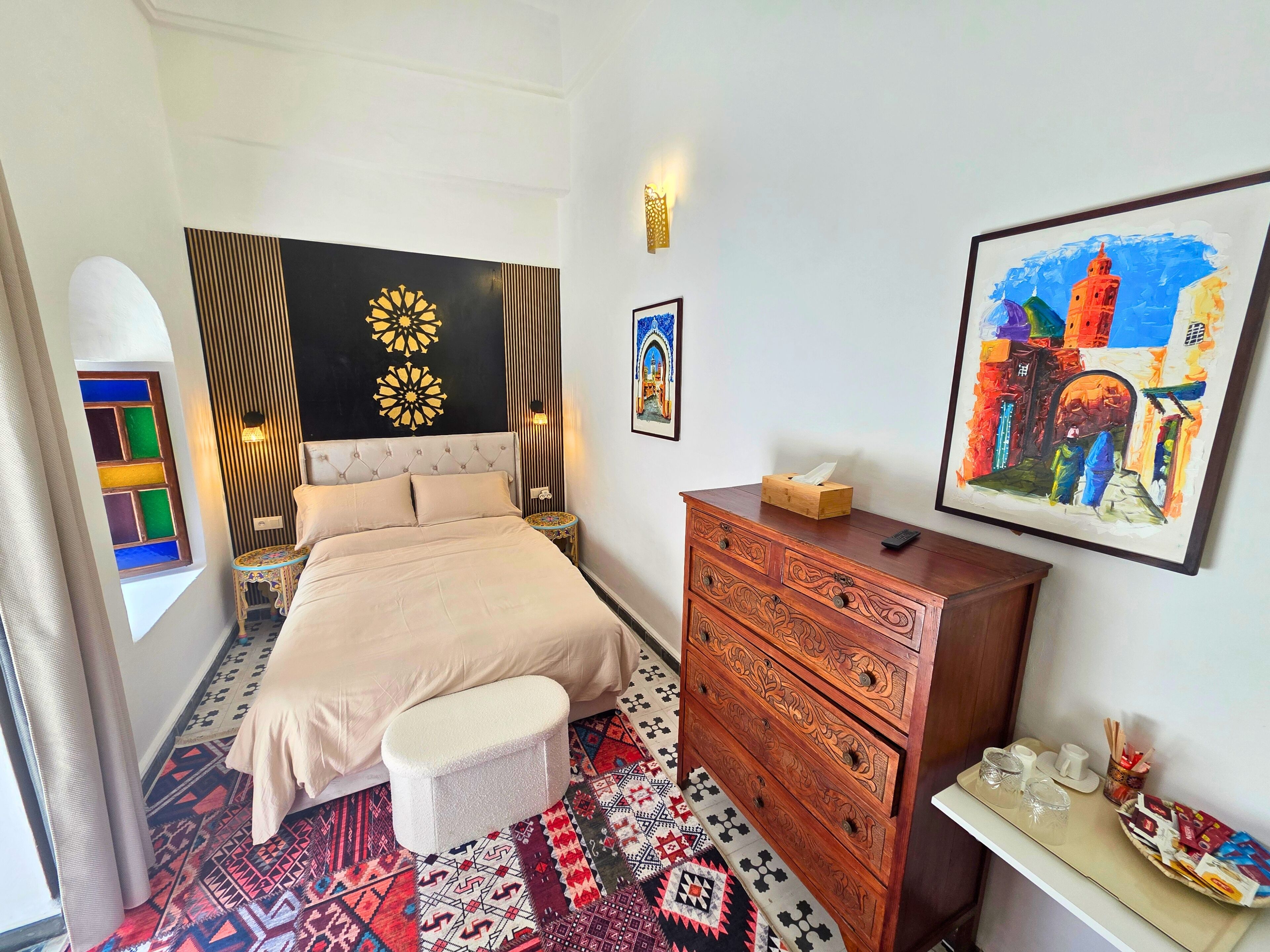A hotel room with a bed, a dresser, a small table with cups and a tray, a window with a colorful view, and a painting on the wall.