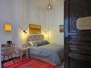 Une chambre à coucher comprenant un lit, une petite table, une chaise, une lampe et une œuvre d’art accrochée au mur.
