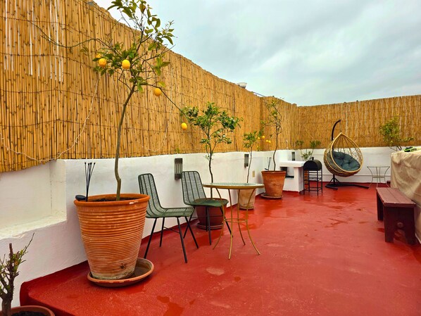 Un espace extérieur aménagé avec des chaises en osier, un fauteuil suspendu et des plantes en pot, le tout adossé à un mur en bambou.