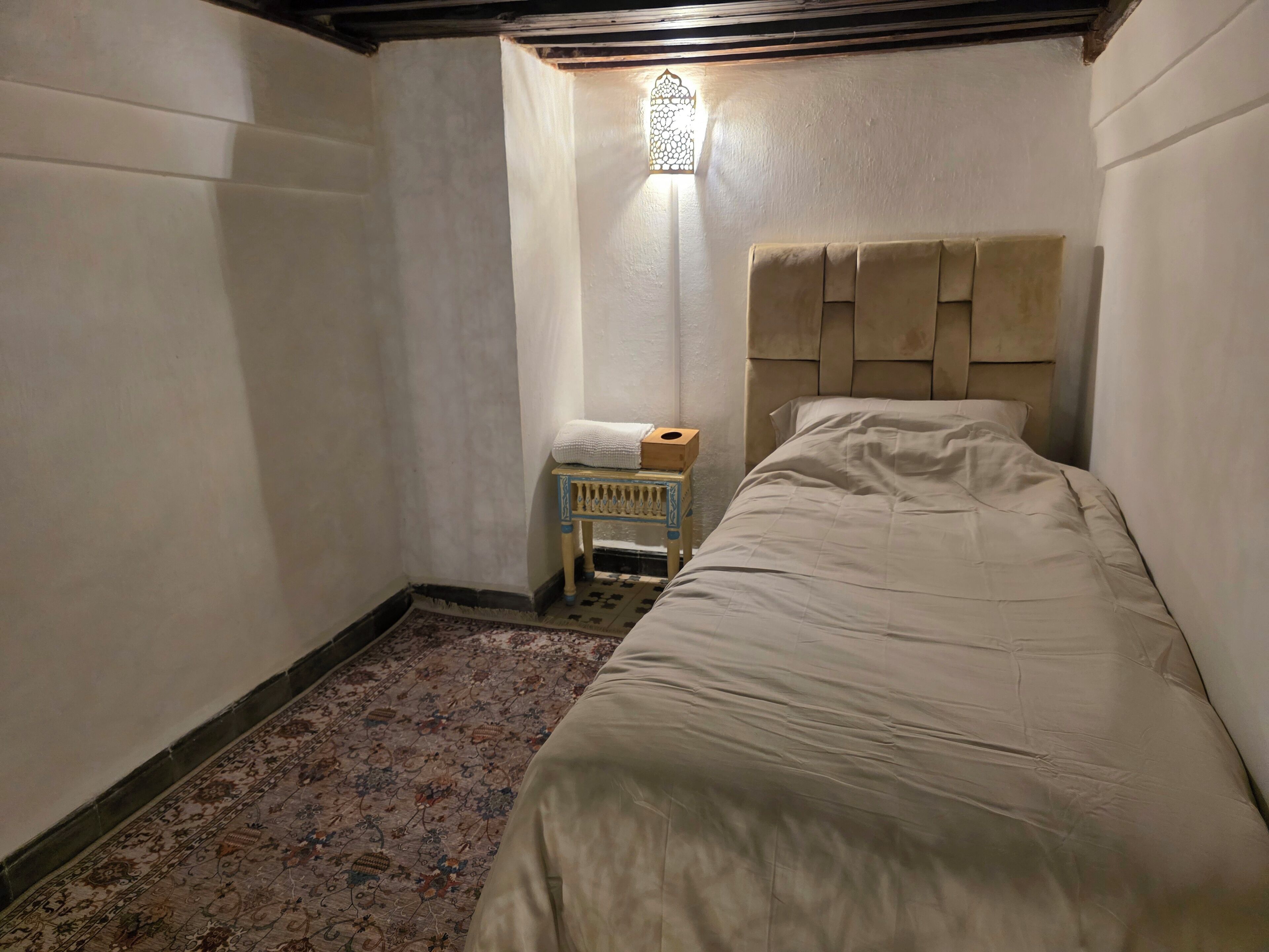 Une petite pièce faiblement éclairée, avec un lit simple, une petite table sur laquelle se trouve une boîte, et un tapis à motifs.