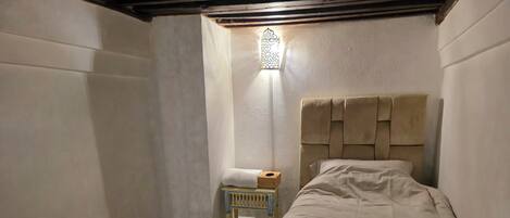 Une petite pièce faiblement éclairée, avec un lit simple, une petite table sur laquelle se trouve une boîte, et un tapis à motifs.