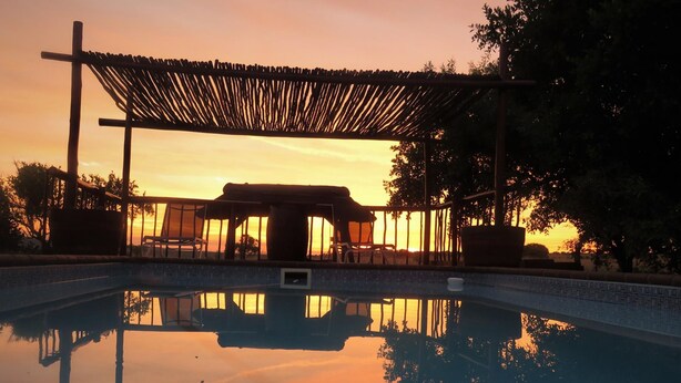Een zwembad met een houten pergola en stoelen, omgeven door bomen, tijdens zonsondergang.