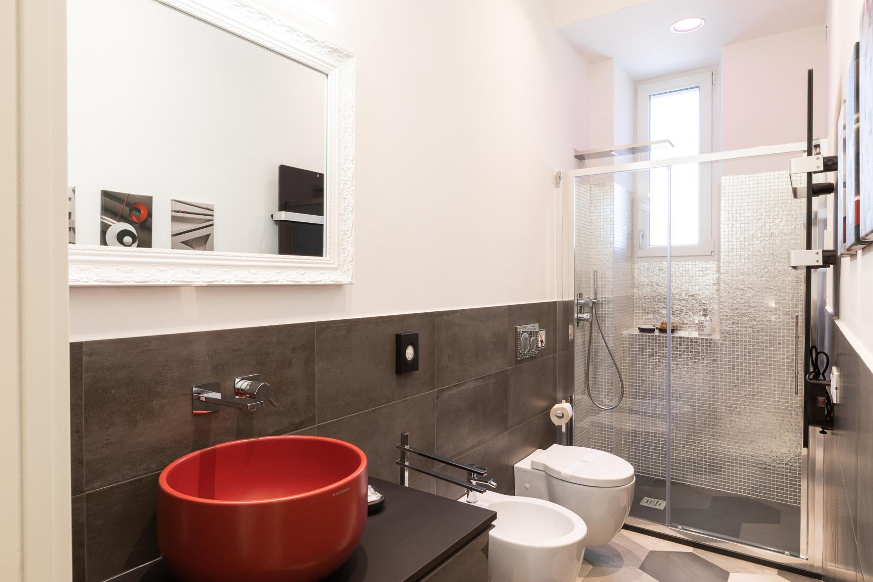 Un baño moderno con un espejo grande, un lavabo rojo, inodoro y cabina de ducha con paredes de cristal.