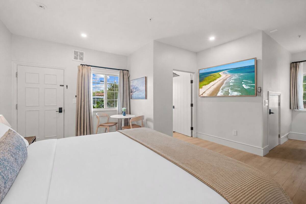 A bedroom with a large bed, a small table with chairs, a painting of a beach, and a door leading to another room.