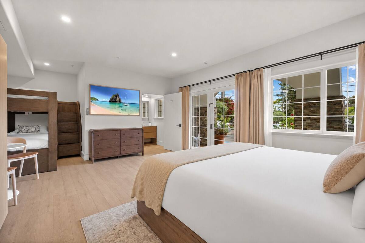 A bedroom with a large bed, a desk, a dresser, and a view of the outdoors through a glass door.