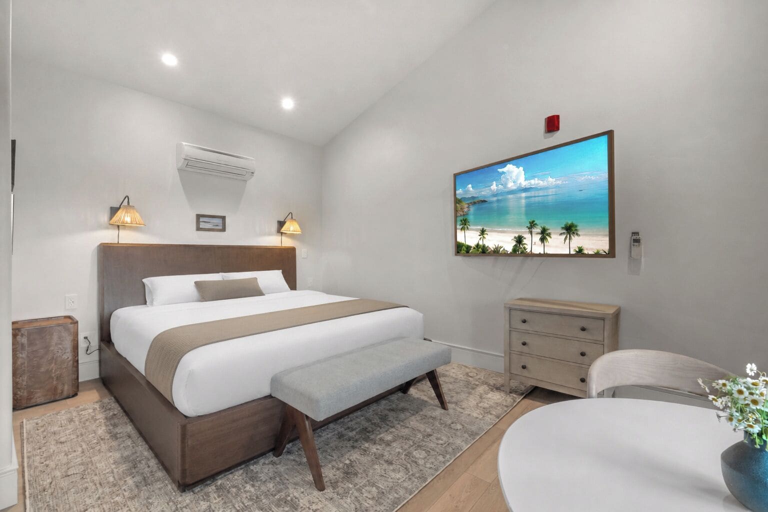 Ein Schlafzimmer mit Bett, Nachttischlampen, einem Nachttisch, einer Kommode, einem Sessel, einem runden Tisch, einem an der Wand befestigten Fernseher und einem Strandbild.