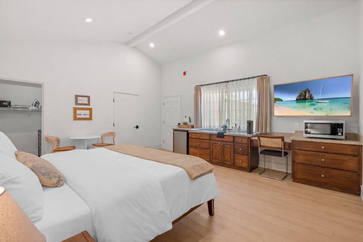 A hotel room with a bed, a desk, a microwave, a sink, and a large window with curtains.