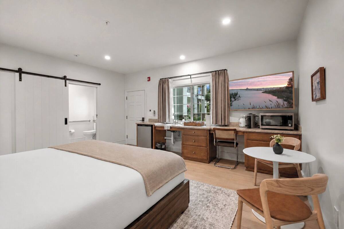 A bedroom with a bed, a desk, a chair, a small table, a microwave, and a painting on the wall.