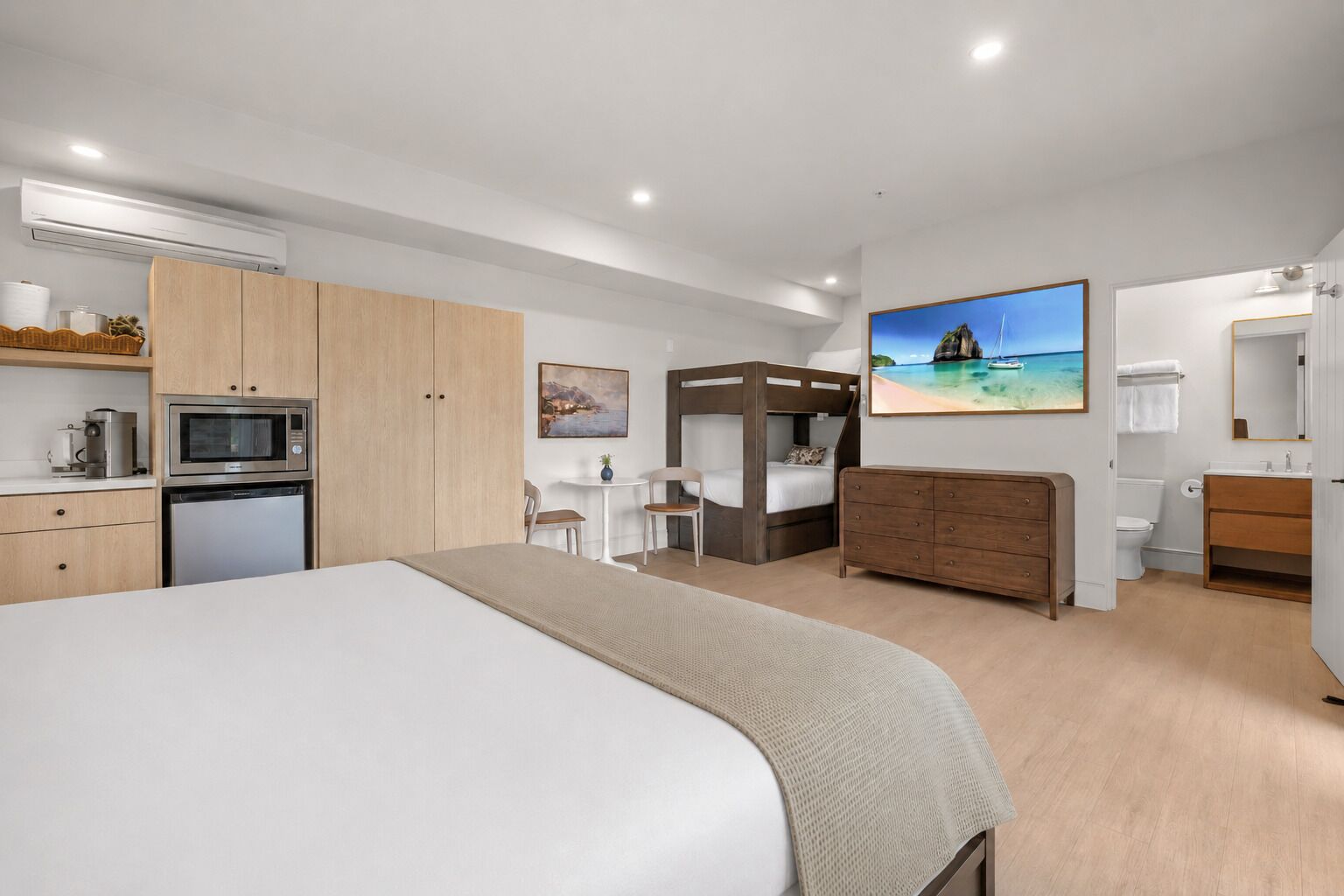 A modern hotel room with a large bed, a kitchenette, a desk, a bunk bed, and a bathroom.