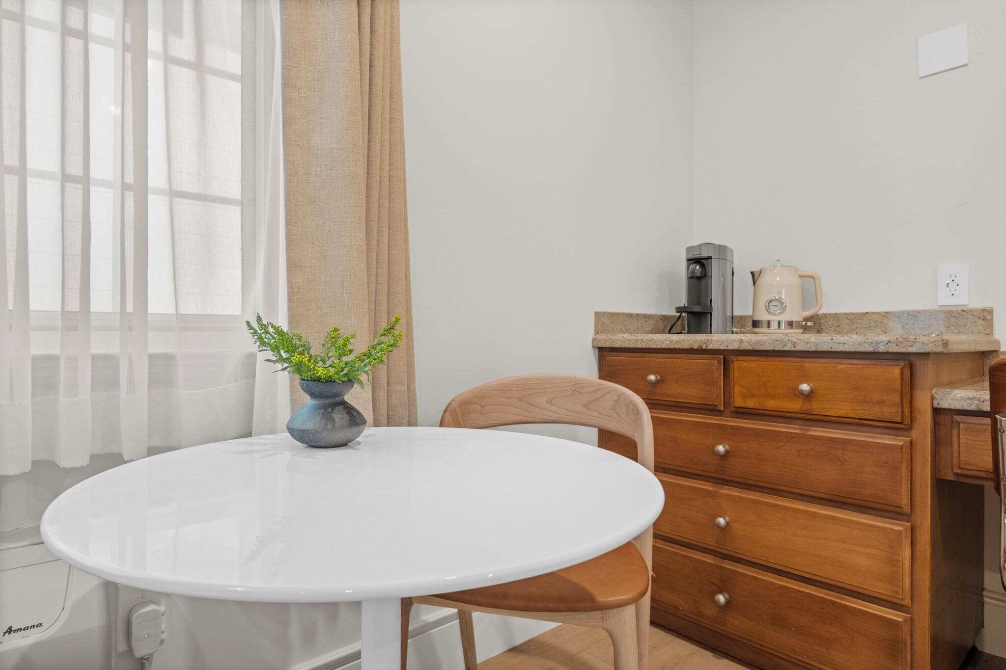 A white round table with a potted plant, a wooden chair, a wooden dresser with drawers, a coffee maker, and a window with sheer curtains.