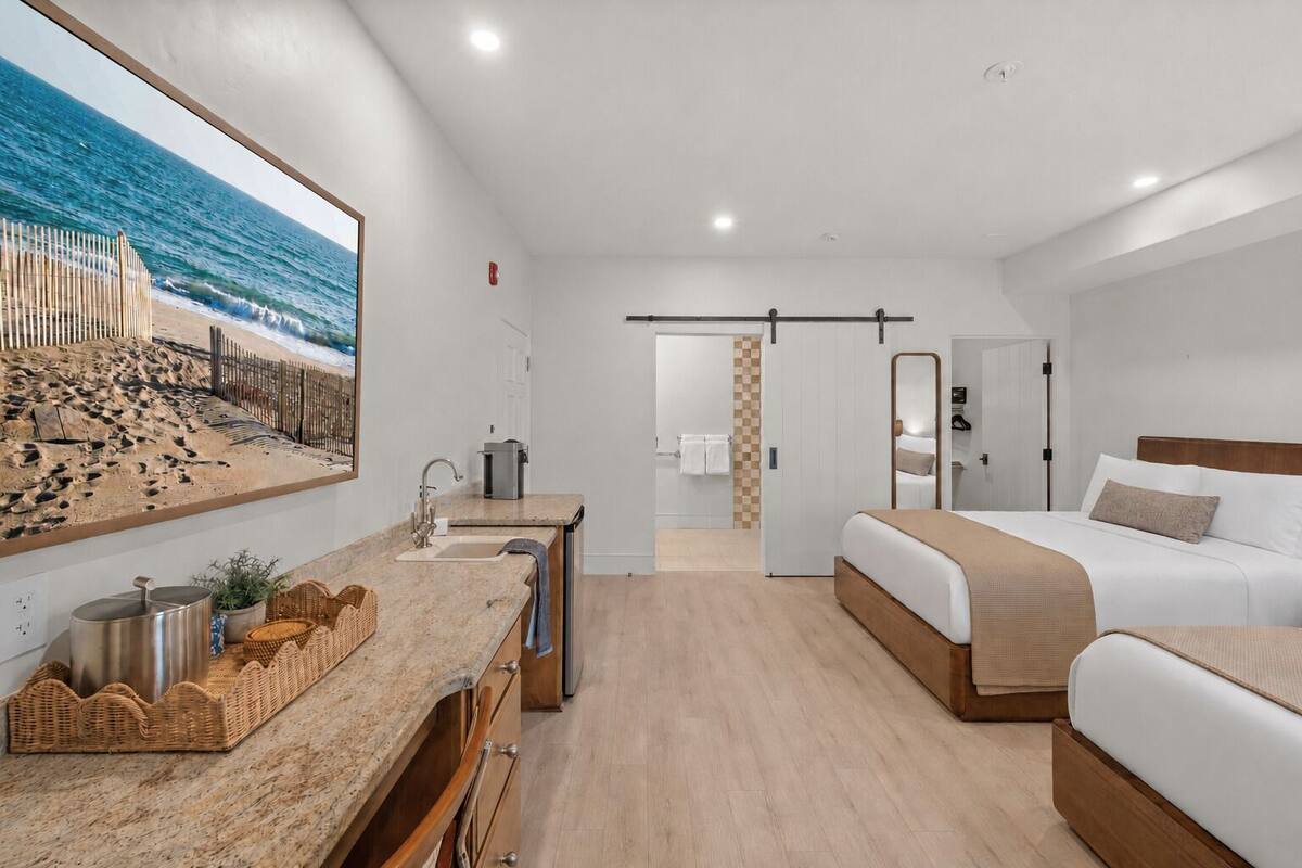 A hotel room with a bed, a desk with a sink, a large beach view painting, and a sliding door leading to the bathroom.