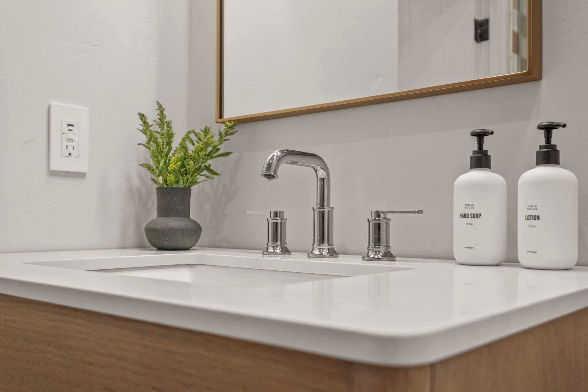 Un baño con un lavabo blanco, un espejo, una planta en maceta y dos botellas de jabón de manos y crema hidratante.