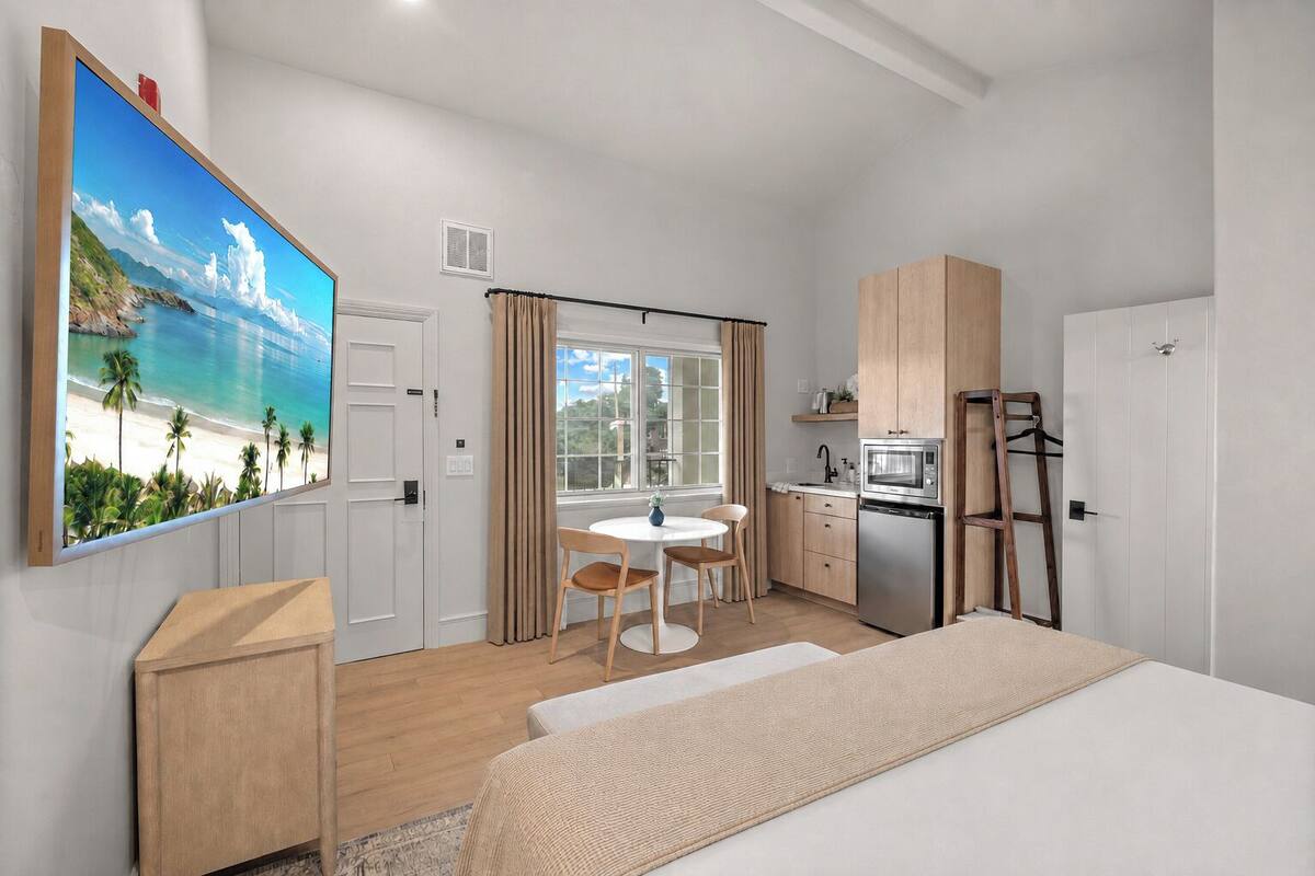 A hotel room with a bed, a dining table with chairs, a kitchenette, and a large window with curtains.