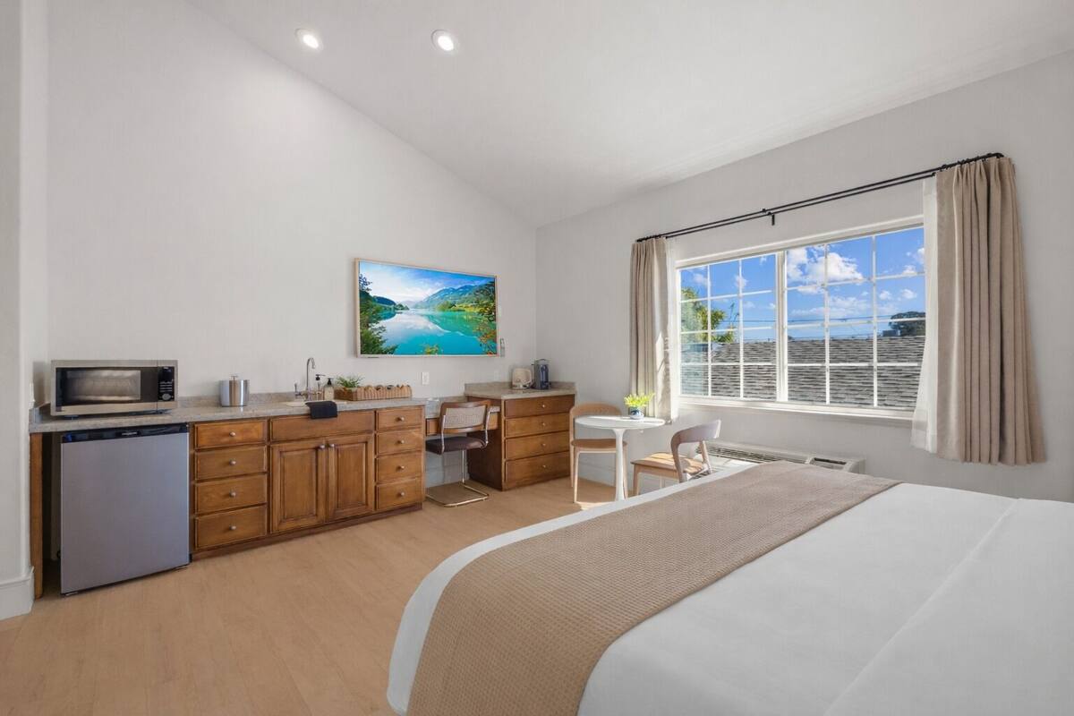 A bedroom with a bed, a desk, a chair, a kitchenette, and a window with a view.