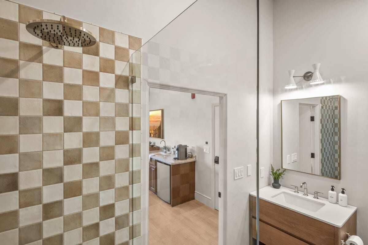 A bathroom with a checkered tile wall, a shower, a sink with a mirror, and a small island with a kettle.