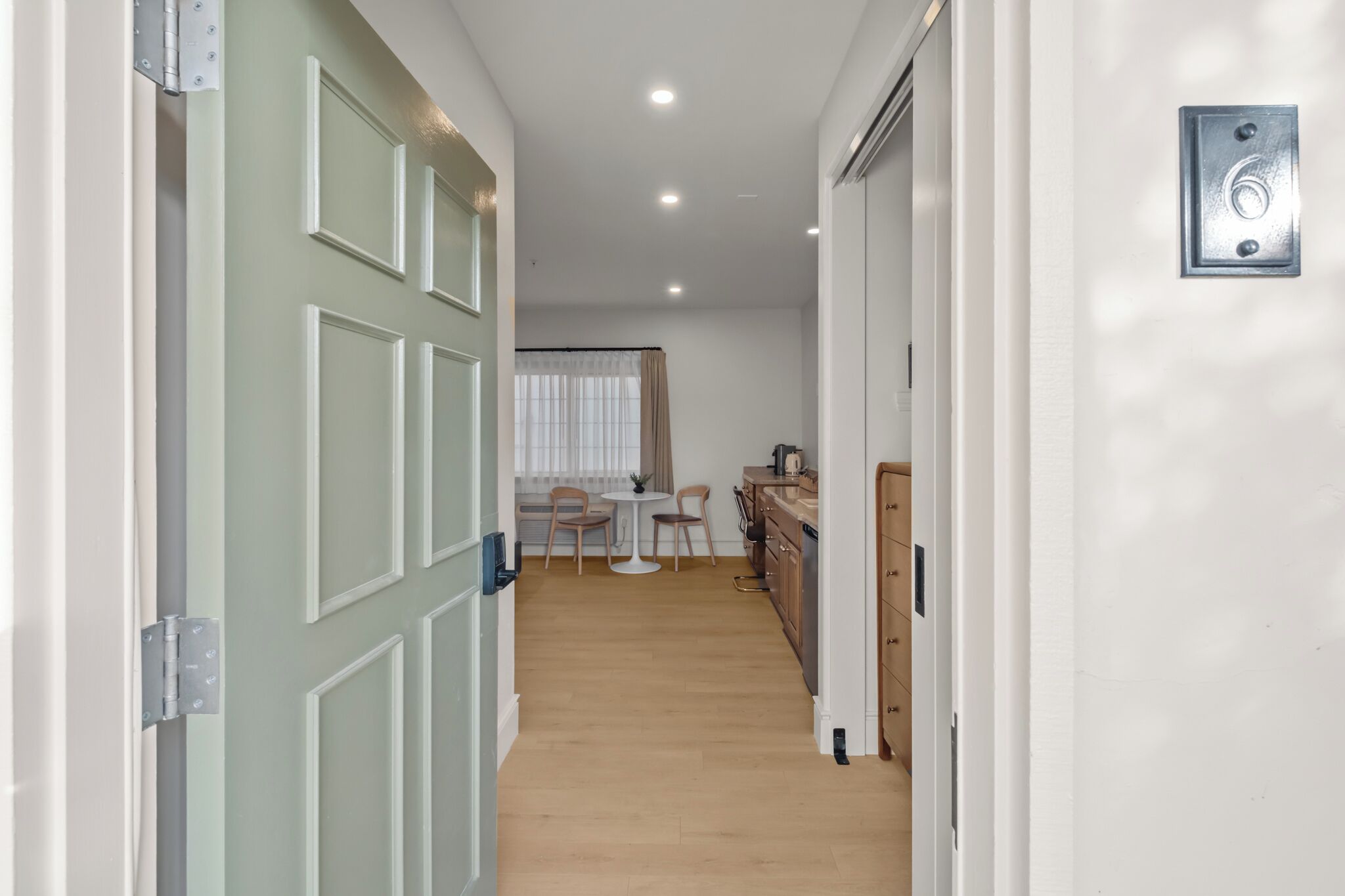A hallway with a door, a small table with chairs, a window with curtains, and a kitchen area.