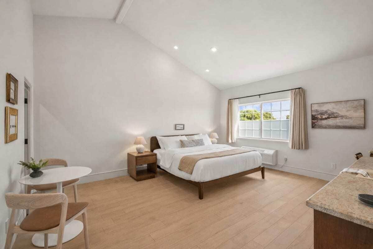 A bedroom with a bed, bedside tables, a small table with chairs, a window with curtains, and a painting on the wall.