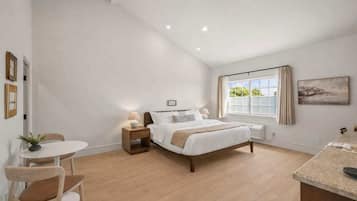 A bedroom with a bed, bedside tables, a small table with chairs, a window with curtains, and a painting on the wall.