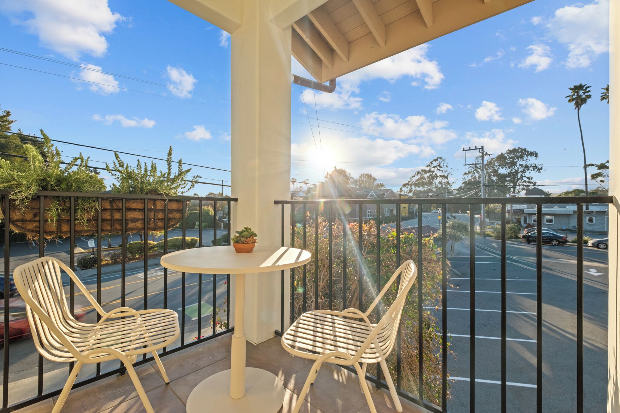 A balcony with two chairs and a table, overlooking a street with parked cars and houses.
