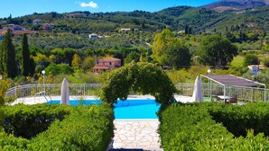 Une piscine entourée de verdure, un sentier en pierre et une vue sur des maisons situées dans une zone vallonnée.