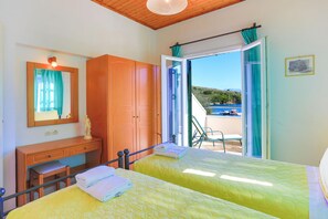 A room with two beds, a wooden wardrobe, a desk, and a mirror. There is a view of the sea through an open door. - Theophilos 1 in Άγιος Στέφανος (Agios Stefanos)