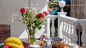 Ein Tisch mit einer Rosenvase, etwas Trockenblume, einer Obstschale und einem Teekännchen auf einem Balkon mit wei ßer Brüstung und Blick auf das Meer.