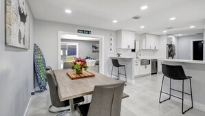Une cuisine moderne avec des meubles blancs, une table à manger en bois agrémentée d’un arrangement floral, et un coin bar avec des tabourets.