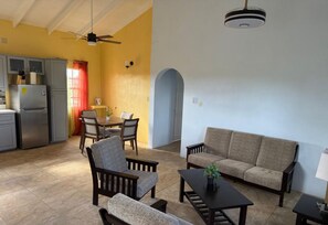 Ein Wohnzimmer mit einer Couch, Sesseln und einem Couchtisch. Es gibt eine Küchenzeile mit Kühlschrank und Schränken. Außerdem ist ein Esstisch mit Stühlen zu sehen.