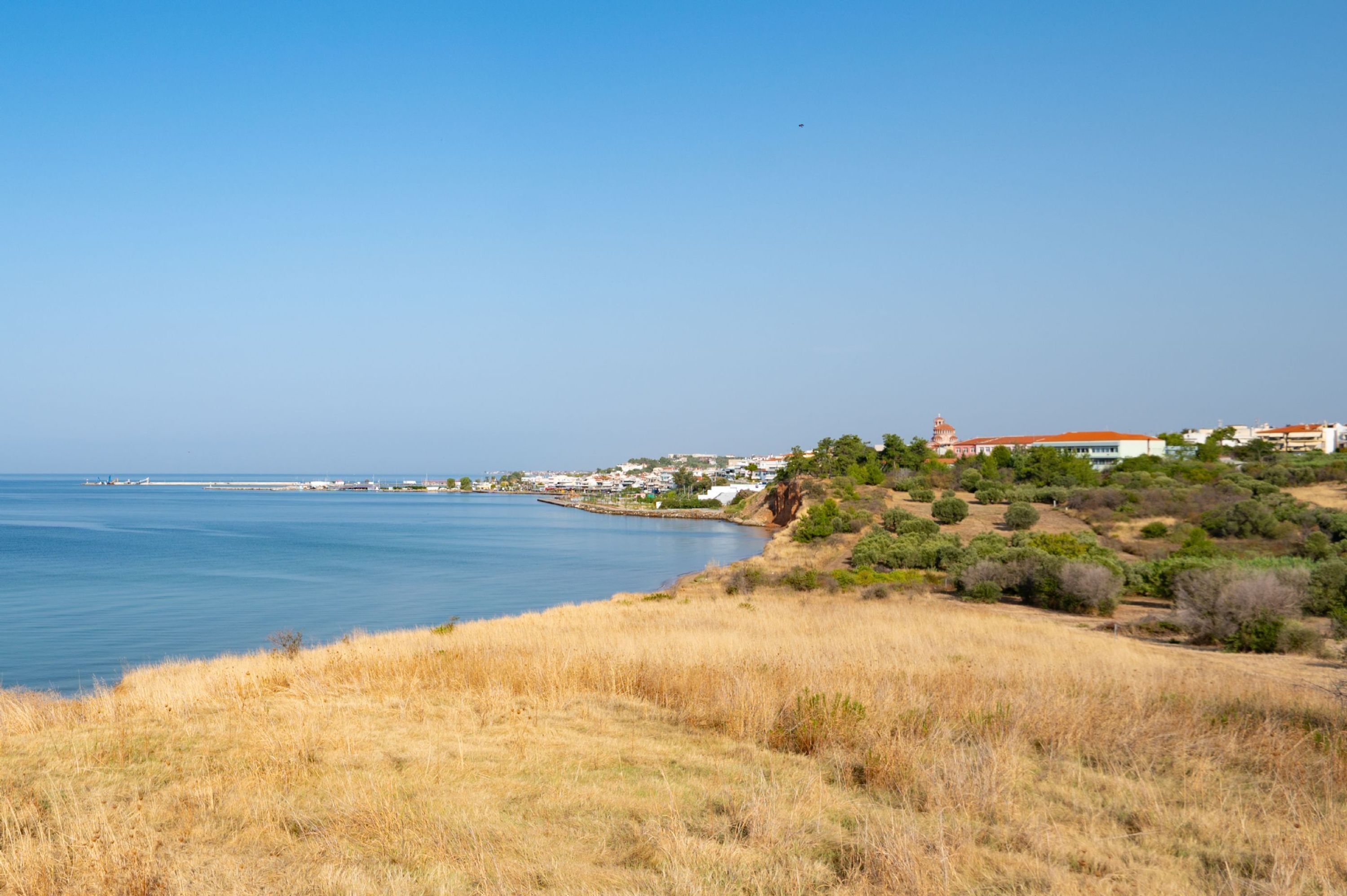 A coastal landscape with a clear sky, a body of water, and a hillside with buildings and vegetation.