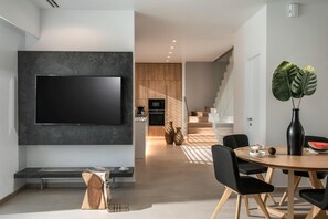 A modern living room with a flat-screen TV mounted on a dark wall, a wooden dining table with black chairs, and a large plant in a black vase. - Made By The Sea Villas 9 BDRM (Mades)