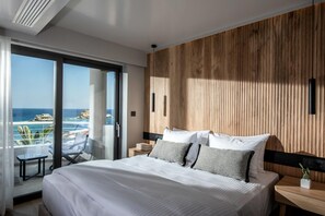 A modern hotel room with a large bed, a wooden accent wall, a balcony with a view of the ocean, and a small table with a chair. - Made By The Sea Villas 9 BDRM (Mades)