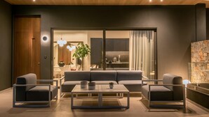 A modern living room with a sofa, two armchairs, and a coffee table. There is a wooden door and a glass wall leading to a kitchen area. - Made By The Sea Villas 8 BDRM (Mades)