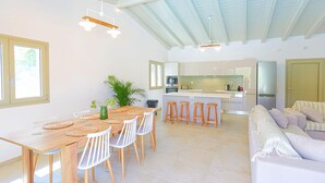 A spacious living area with a wooden dining table, white chairs, and a sofa. A kitchen with modern appliances and a high ceiling is visible in the background. - Villa Jupeter in Corfu (Corfu)