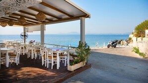 A covered outdoor seating area with white tables and chairs, overlooking a beach and the sea. - Takis House in Katavolos (Katavolos)