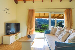 A living room with a TV on a wooden stand, a sofa with patterned cushions, and a view of a garden through large windows. - Green Family Villa in Agios Ioannis (Agios Ioannis)