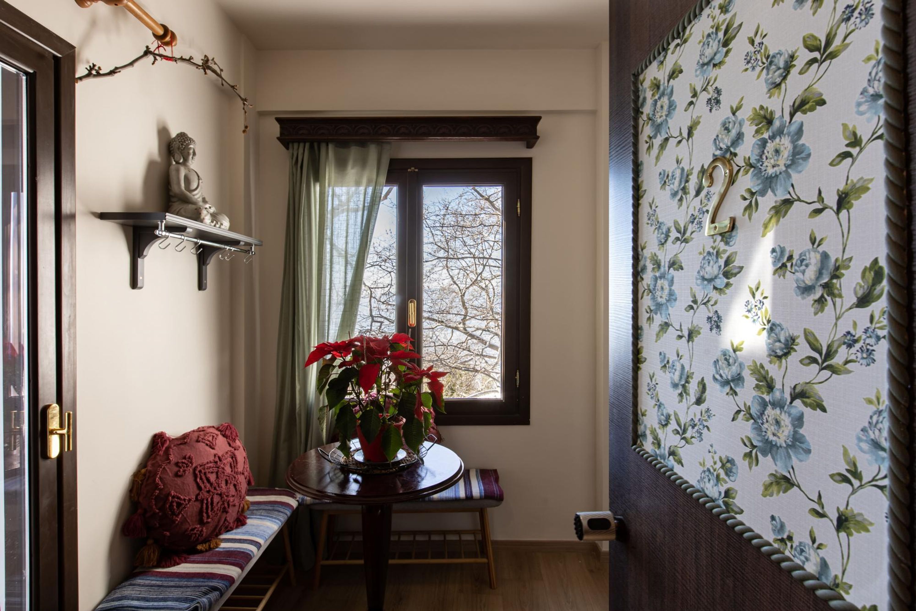 Ein Zimmer mit einer hölzernen Bank, einem kleinen Tisch mit einem Topfpflanzen, einer Buddha-Statue auf einem Wandregal und einem Fenster mit Blick auf kahle Bäume.