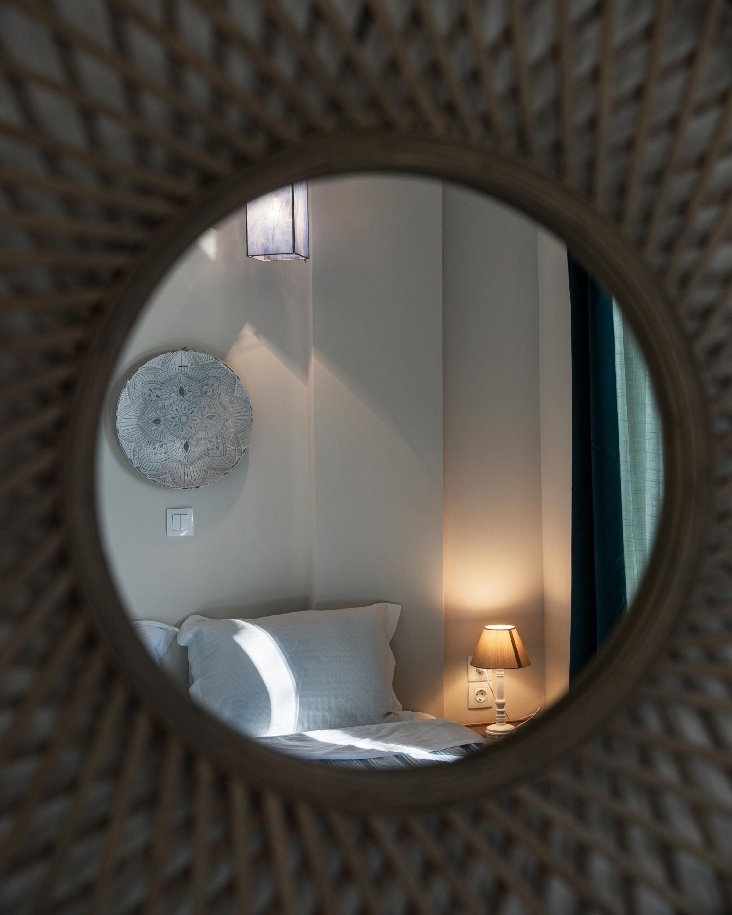 En rund spegel som reflekterar en säng med en vit kudde, en väggmonterad lampa och ett sängbord med en lampa.