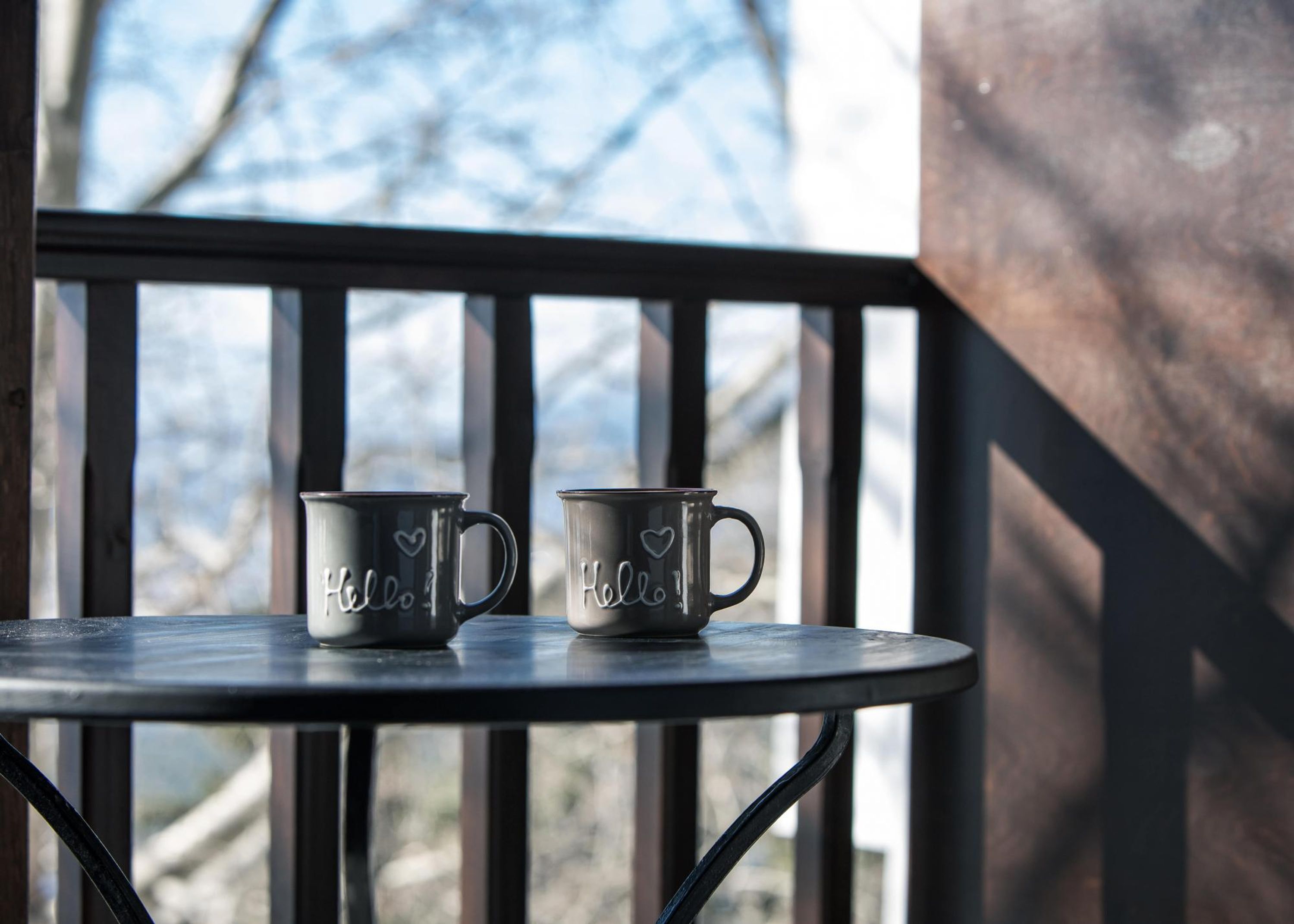 Deux tasses en métal ornées des mots « Bonjour » et de motifs en forme de cœur, posées sur une table en métal sur un balcon.