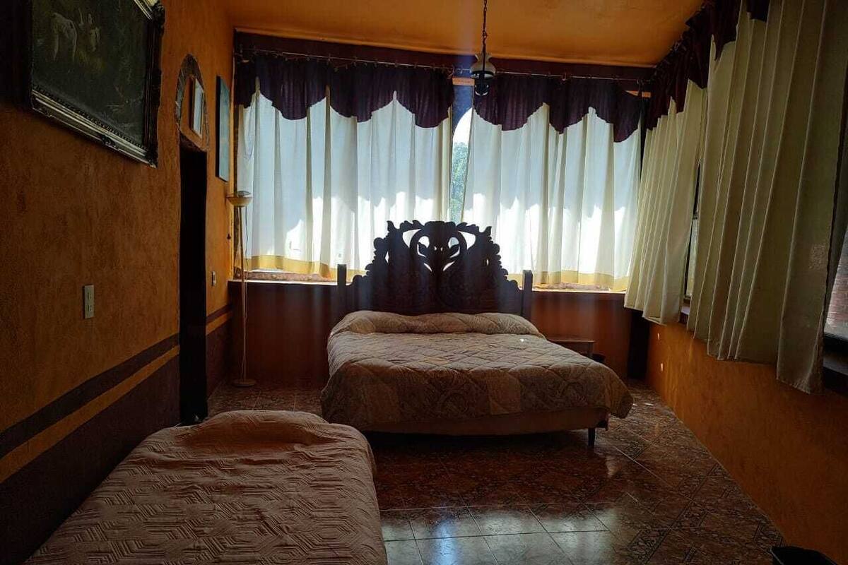 A room with two beds, a wooden headboard, a window with curtains, and a patterned carpet.