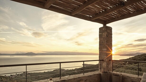 Une terrasse offrant une vue sur la mer et les montagnes au coucher du soleil, avec un pilier en pierre, des chaises en osier et un canapé.