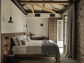 Une chambre à coucher avec une tête de lit en bois, un lit recouvert d’une couverture à motifs, une table de chevet avec une lampe, et une vue sur un mur en pierre par la fenêtre.