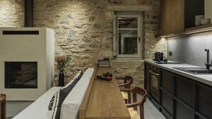 A rustic kitchen with a stone wall, wooden beams, a long wooden table, and a built-in stove. - Aphrodite Suite by Peliva Nature & Suites (Pinakates)