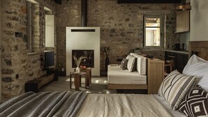 A rustic room with exposed wooden beams, a stone wall, a fireplace, a sofa, a bed, and a small table with a vase. - Aphrodite Suite by Peliva Nature & Suites (Pinakates)