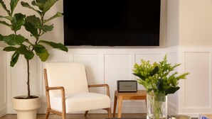 Ein Wohnzimmer mit einem weißen Sessel, einem kleinen Tisch mit einem Radio, einer Topfpflanze und einem an der Wand befestigten Fernseher.
