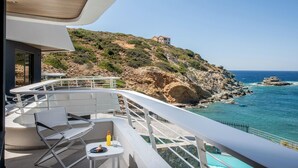 Un balcón con mobiliario blanco, una pequeña mesa con una bebida y vistas al mar y una ladera rocosa.