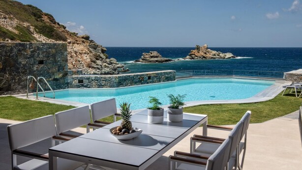 Una piscina con vistas al mar y acantilados rocosos, rodeada por un patio con sillas y una mesa.