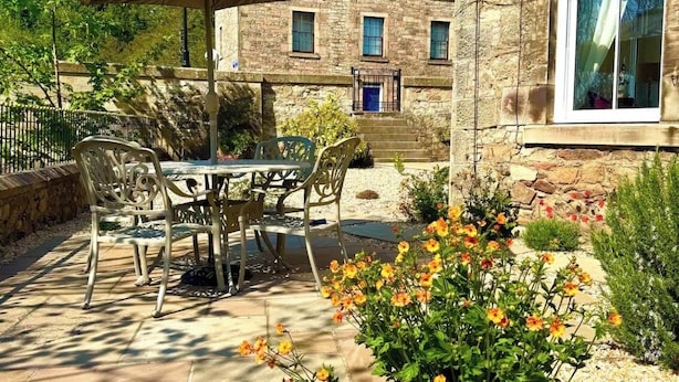 Un patio al aire libre con una mesa y sillas, rodeado de un jardín con flores y un edificio de piedra al fondo.