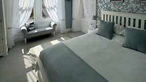 Un dormitorio con una cama grande, una mesita de noche, un banco y una ventana con cortinas ligeras.