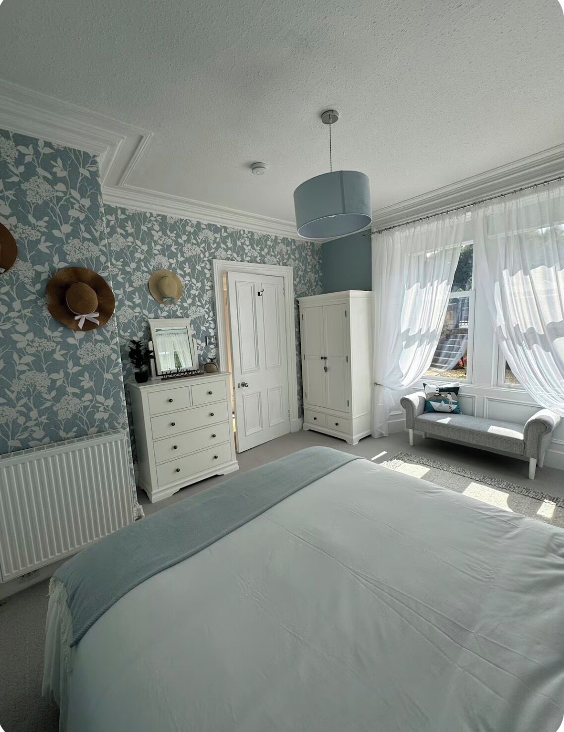 A bedroom with a large bed, a dresser, a wardrobe, a vanity with a mirror, and a window with sheer curtains.